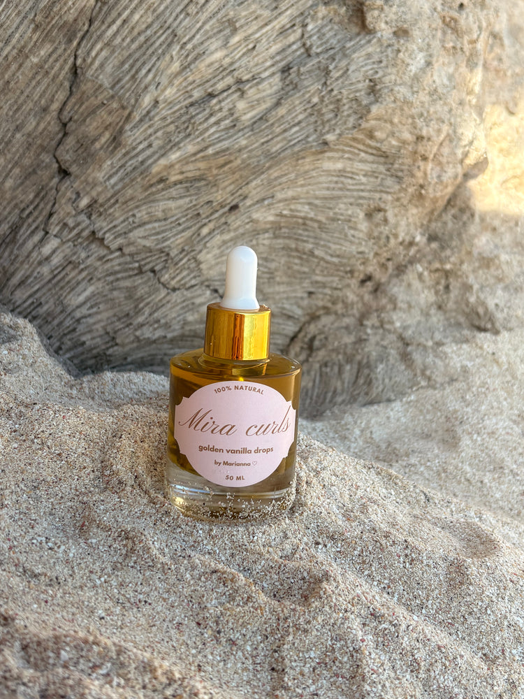 Mira Curls golden vanilla drops nestled in sand against driftwood